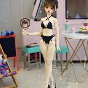 May include: A doll wearing a black bikini and black high heels stands on a brown base. The doll has a bun hairstyle and is posed in front of a pink backdrop with a small table and a miniature food display.