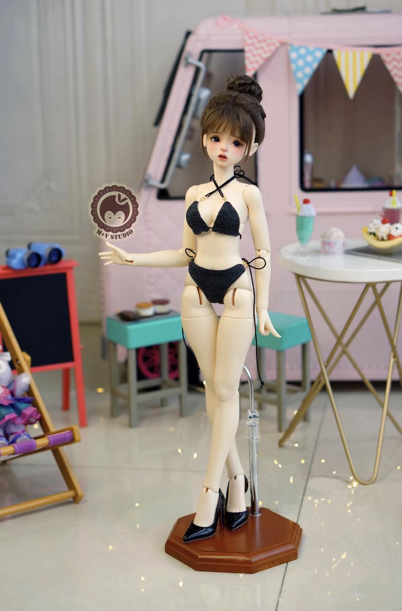 May include: A doll wearing a black bikini and black high heels stands on a brown base. The doll has a bun hairstyle and is posed in front of a pink backdrop with a small table and a miniature food display.