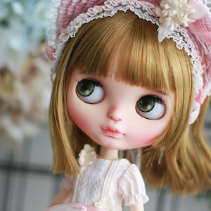 May include: A doll with blonde hair and green eyes wearing a pink and white lace hat and a white dress with a pink ribbon. The doll is looking to the left and has a sweet expression on her face. The background is blurred and out of focus.