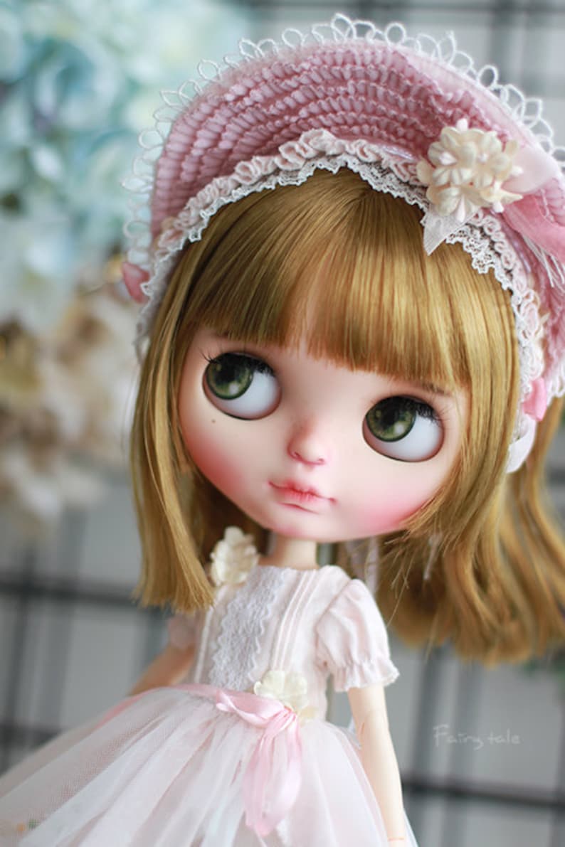 May include: A doll with blonde hair and green eyes wearing a pink and white lace hat and a white dress with a pink ribbon. The doll is looking to the left and has a sweet expression on her face. The background is blurred and out of focus.