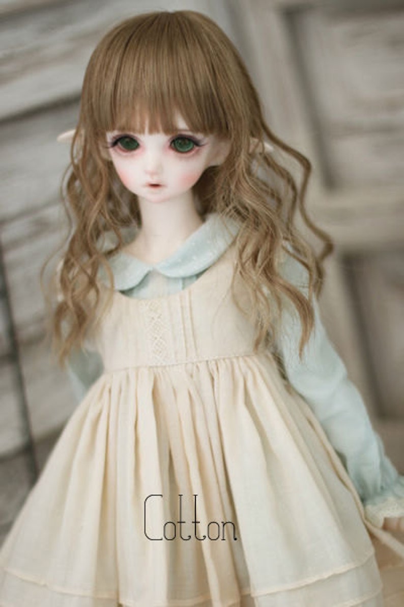 May include: A doll with long, wavy brown hair wearing a white dress with a blue collar and the word "Cotton" printed on the front.
