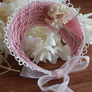 May include: A pink and white crocheted hat with a lace trim and a bow. The hat is decorated with a cluster of white flowers.