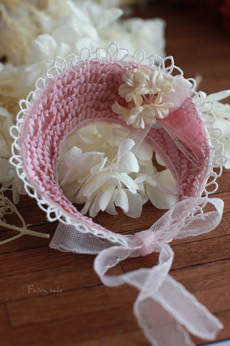 May include: A pink and white crocheted hat with a lace trim and a bow. The hat is decorated with a cluster of white flowers.