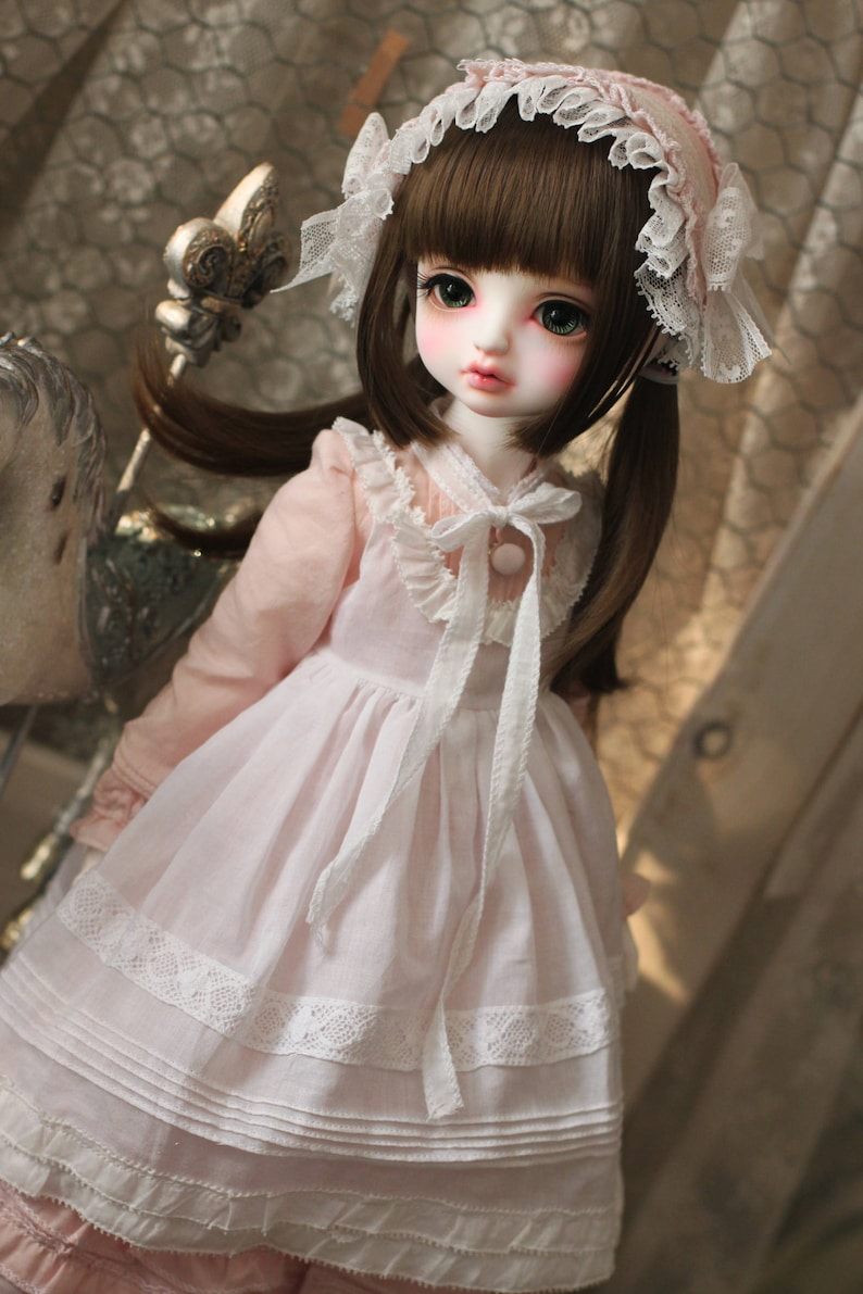 May include: A porcelain doll with brown hair, wearing a pink and white dress with lace trim and a white lace headband. The doll has green eyes and a pink blush on her cheeks.