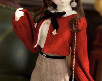 May include: A doll wearing a red knitted cardigan with a white trim, a black velvet bow, and a brown knitted skirt. The doll is also wearing black tights with a white lace trim and a black purse with a gold chain.
