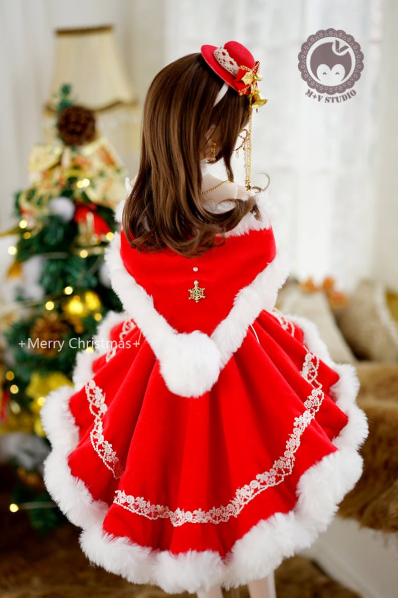 May include: A red velvet dress with white fur trim and a white lace overlay. The dress has a hood and a snowflake brooch. The dress is worn by a doll with long brown hair and a red hat with a white bow.