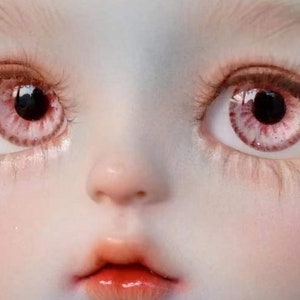 Op de afbeelding: Close-up van het gezicht van een pop met roze en witte ogen. De pop heeft een lichtroze huidskleur en een kleine, licht geopende mond.