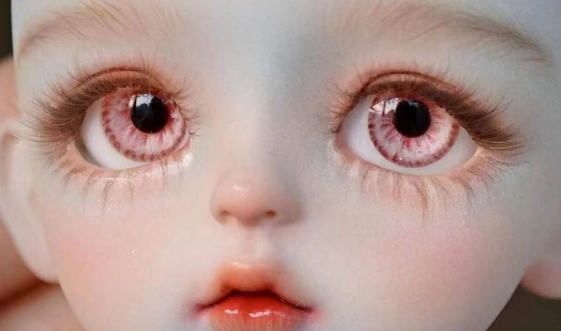 Op de afbeelding: Close-up van het gezicht van een pop met roze en witte ogen. De pop heeft een lichtroze huidskleur en een kleine, licht geopende mond.