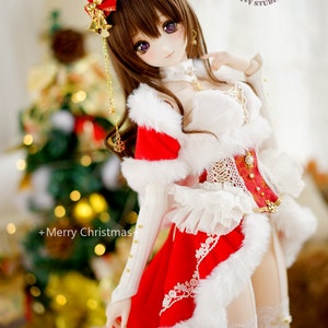 May include: A doll wearing a red and white Christmas outfit with a white fur trim. The doll has long brown hair, purple eyes, and a red hat with gold bells. The doll is standing in front of a Christmas tree with lights.