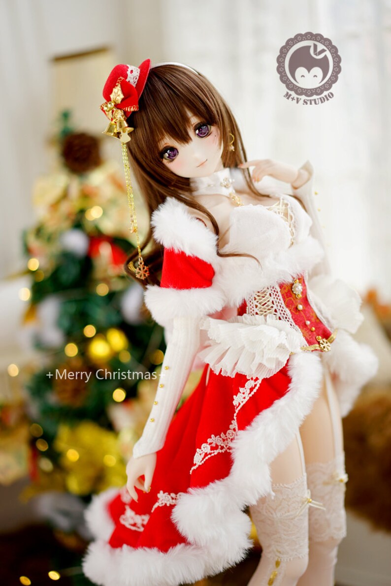 May include: A doll wearing a red and white Christmas outfit with a white fur trim. The doll has long brown hair, purple eyes, and a red hat with gold bells. The doll is standing in front of a Christmas tree with lights.