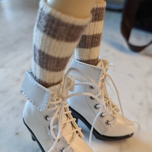 Puede incluir: Botas blancas de tobillo con calcetines a rayas marrones y blancas. Las botas tienen cordones y una punta puntiaguda.