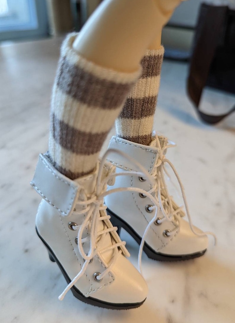 Puede incluir: Botas blancas de tobillo con calcetines a rayas marrones y blancas. Las botas tienen cordones y una punta puntiaguda.