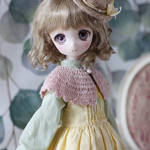 May include: A doll with light brown curly hair wearing a yellow dress with a pink crocheted collar and a brown straw hat with a yellow ribbon.