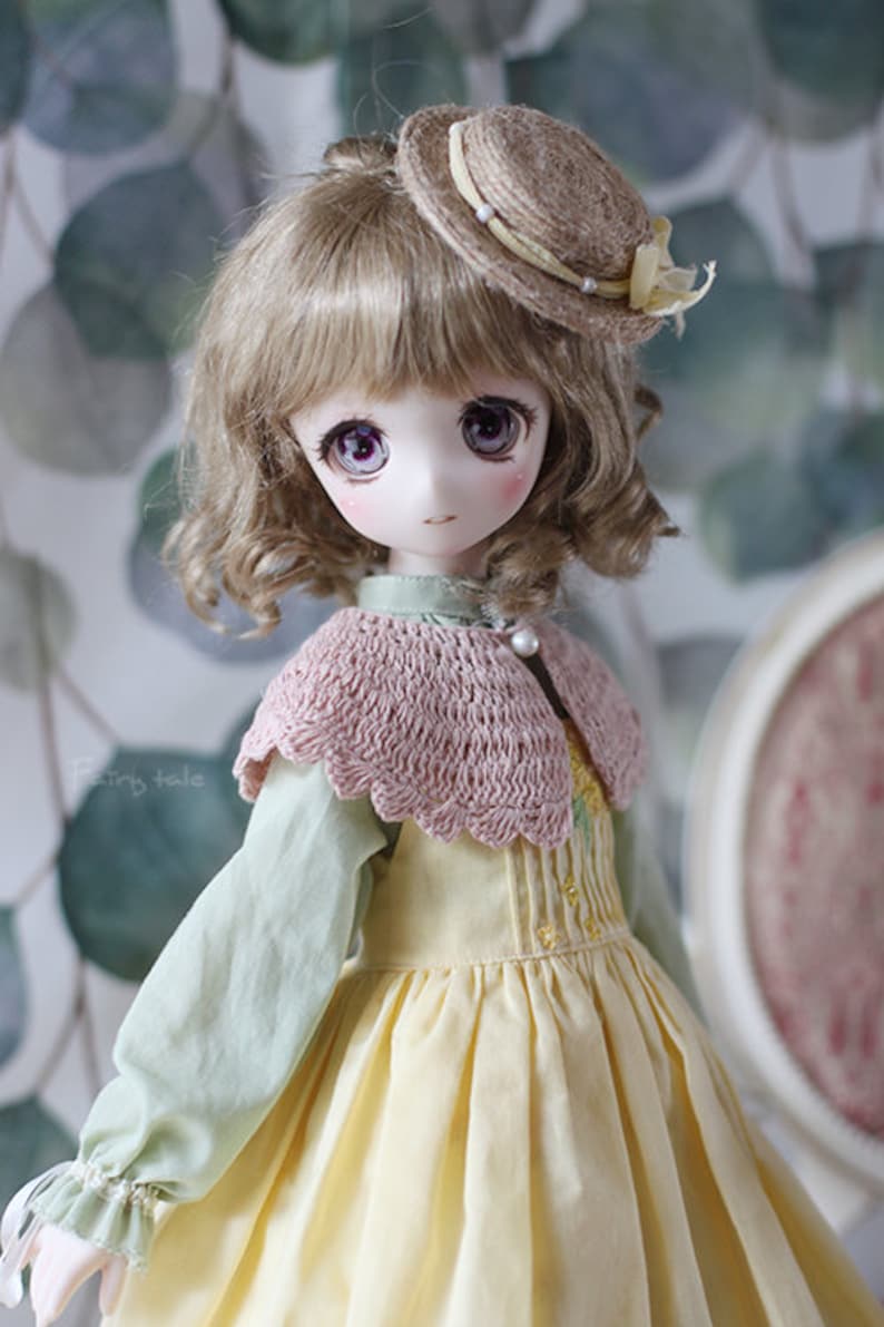 May include: A doll with light brown curly hair wearing a yellow dress with a pink crocheted collar and a brown straw hat with a yellow ribbon.