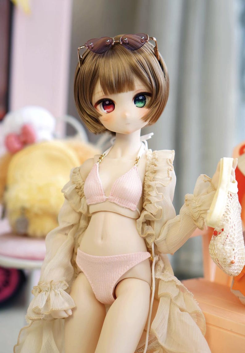 May include: A doll with short brown hair, wearing a pink bikini, a sheer beige robe, and sunglasses. The doll has heterochromia, with one red and one green eye. It holds a small, white, crocheted item.