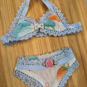 Könnte beinhalten: Ein blaues und weißes Dessous-Set mit Blumenmuster und silbernem Besatz. Das Oberteil ist im Halstert-Stil mit einer Schleife vorne. Das Unterteil ist im Bikini-Stil mit einer Blumenverzierung.