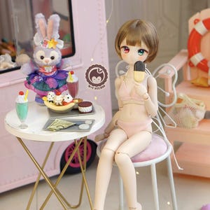 May include: A doll in a pink bikini holding an ice cream bar, seated on a small stool. A table with desserts and a stuffed animal are nearby. The scene is set in front of a pink ice cream truck.