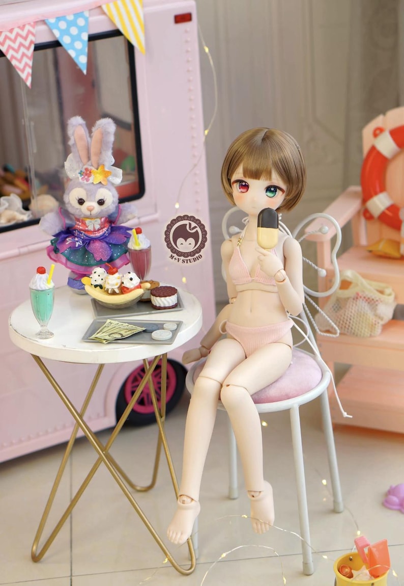 May include: A doll in a pink bikini holding an ice cream bar, seated on a small stool. A table with desserts and a stuffed animal are nearby. The scene is set in front of a pink ice cream truck.