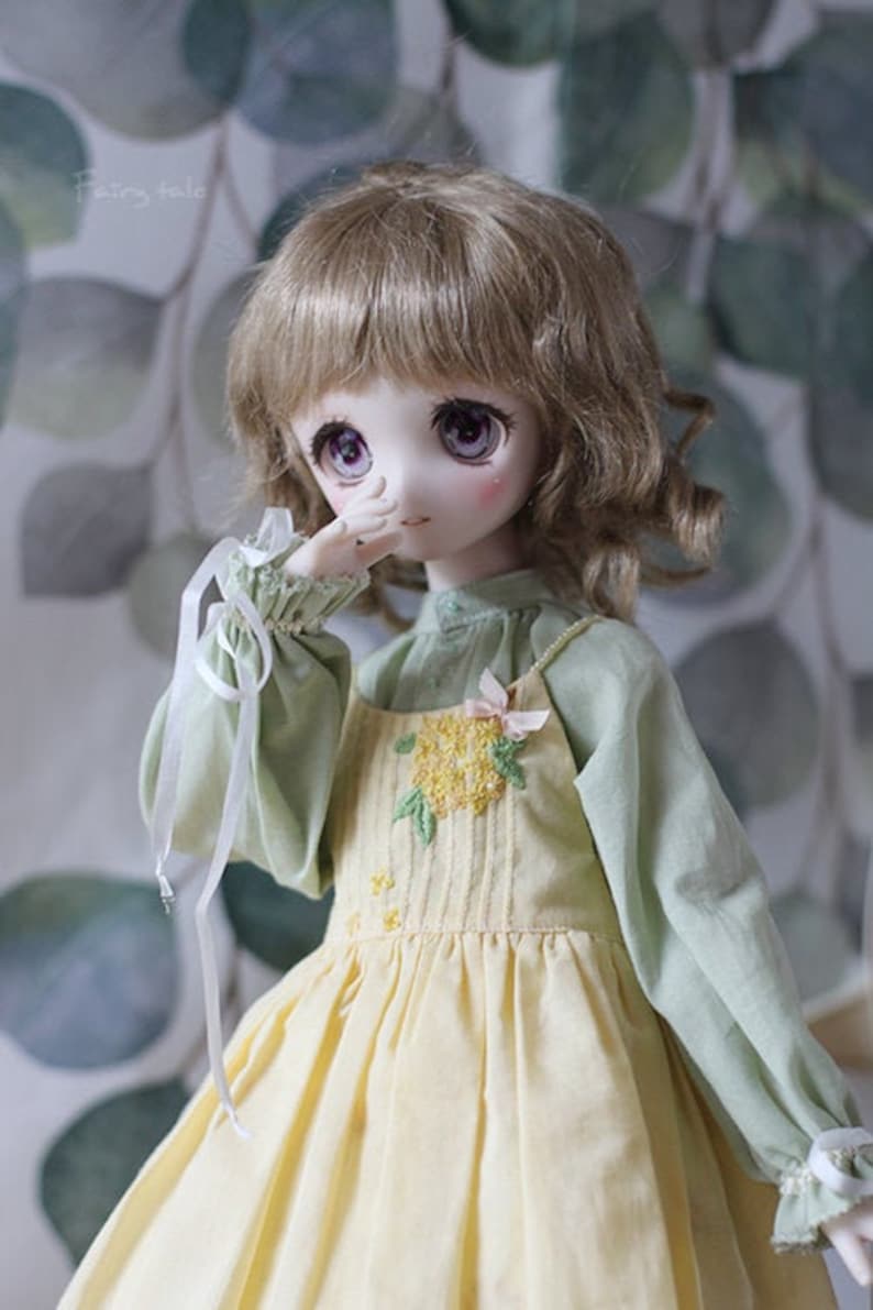 May include: A doll with light brown curly hair wearing a yellow dress with a floral design and a green shirt with white lace trim.