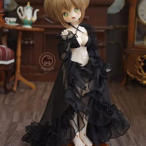 May include: A doll with light brown hair styled in two buns, green eyes, and pale skin. The doll is wearing a black lingerie set with a sheer, ruffled overdress. The doll is standing on a wooden stand in a room with antique furniture.
