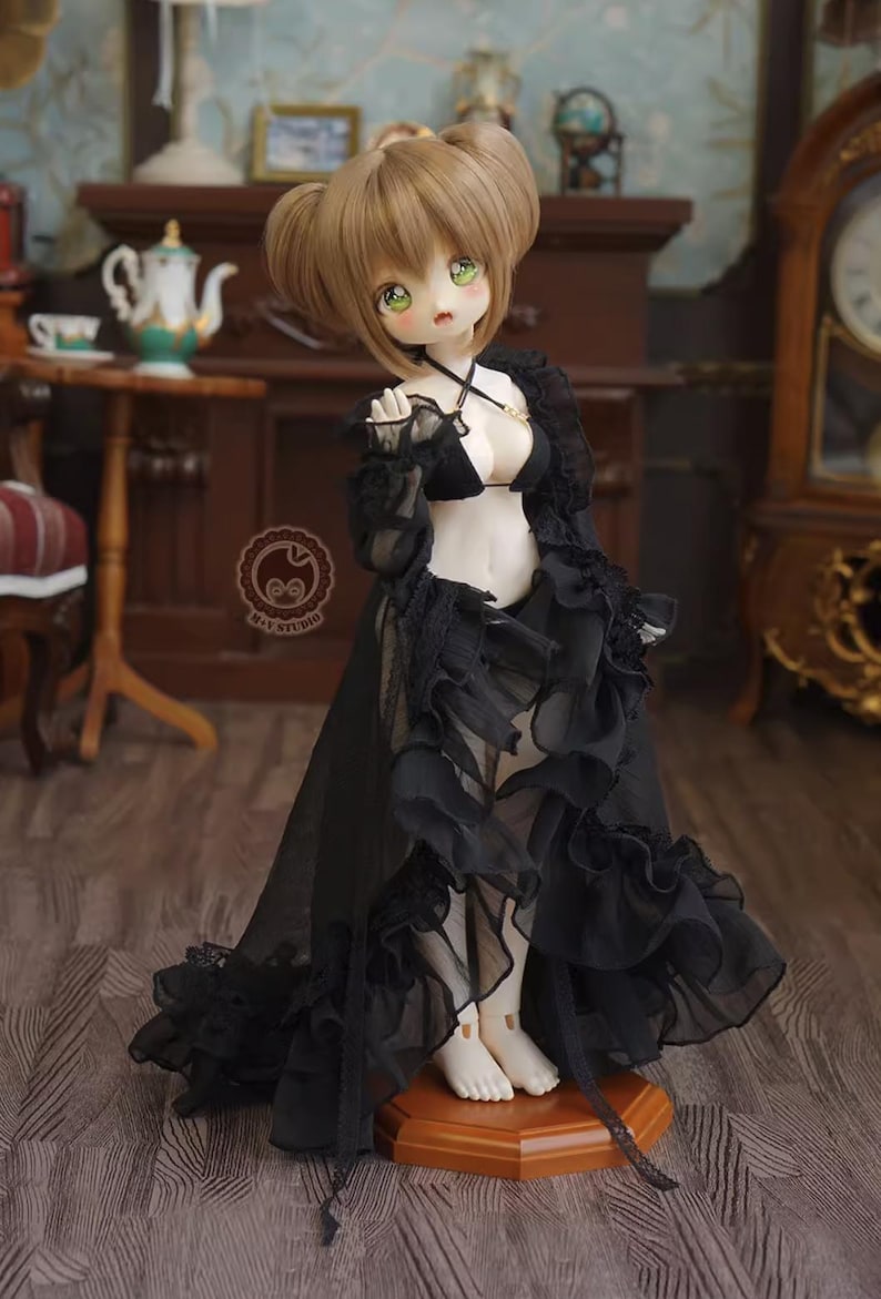 May include: A doll with light brown hair styled in two buns, green eyes, and pale skin. The doll is wearing a black lingerie set with a sheer, ruffled overdress. The doll is standing on a wooden stand in a room with antique furniture.
