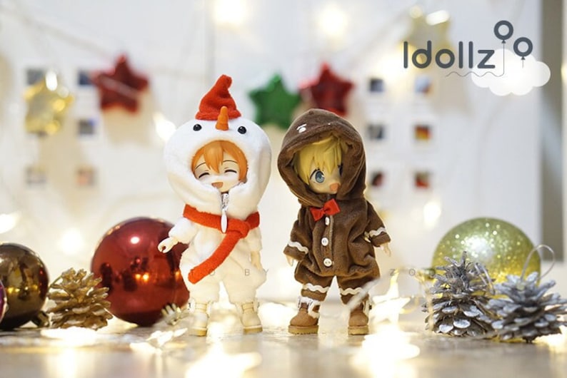 Puede incluir: Dos mu&ntilde;ecas peque&ntilde;as que llevan atuendos navide&ntilde;os. Una mu&ntilde;eca lleva un disfraz de mu&ntilde;eco de nieve blanco con una bufanda roja. La otra mu&ntilde;eca lleva un disfraz de hombre de jengibre marr&oacute;n con una corbata de lazo roja. Las mu&ntilde;ecas est&aacute;n de pie sobre una mesa con decoraciones navide&ntilde;as.