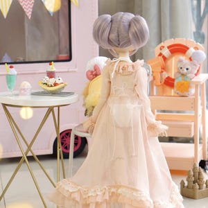 May include: A doll with lavender hair in a light pink dress with lace trim. The doll is standing near a pink ice cream truck and a small table with desserts. A small lifeguard stand with a stuffed animal is in the background.