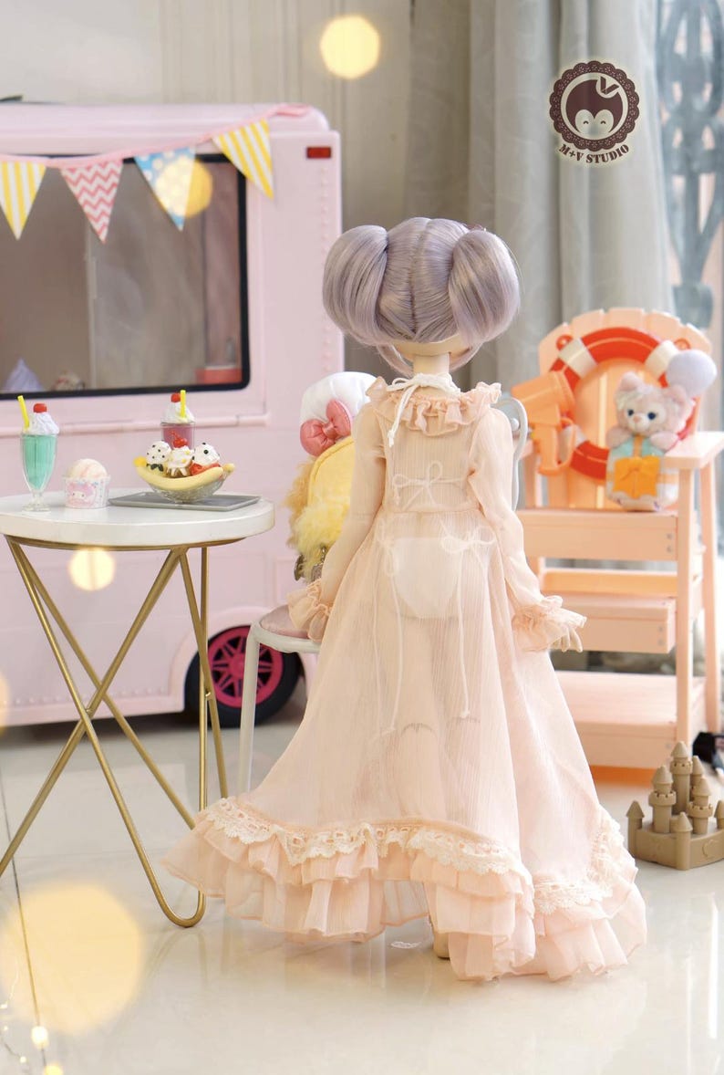 May include: A doll with lavender hair in a light pink dress with lace trim. The doll is standing near a pink ice cream truck and a small table with desserts. A small lifeguard stand with a stuffed animal is in the background.