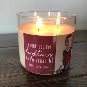 May include: A glass candle jar with a red label that says "Thank you for lighting up our child's life. Love, The McAllisters." The label also has a photo of a young boy wearing a red sweater vest. The candle is lit and the flames are visible.