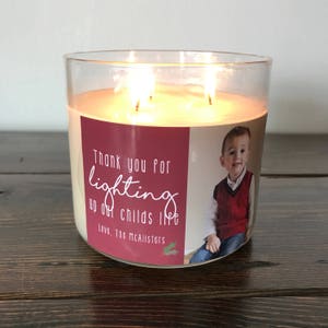 May include: A clear glass candle jar with a red and white label. The label has a photo of a young boy and the text "Thank you for lighting up our childs life Love, The McAllisters".