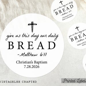 May include: White round labels with black text. The labels feature a cross and the words "give us this day our daily BREAD - Matthew 6:11." One label reads "Christian's Baptism 7.28.2026." Another says "Christian's First Holy Communion."