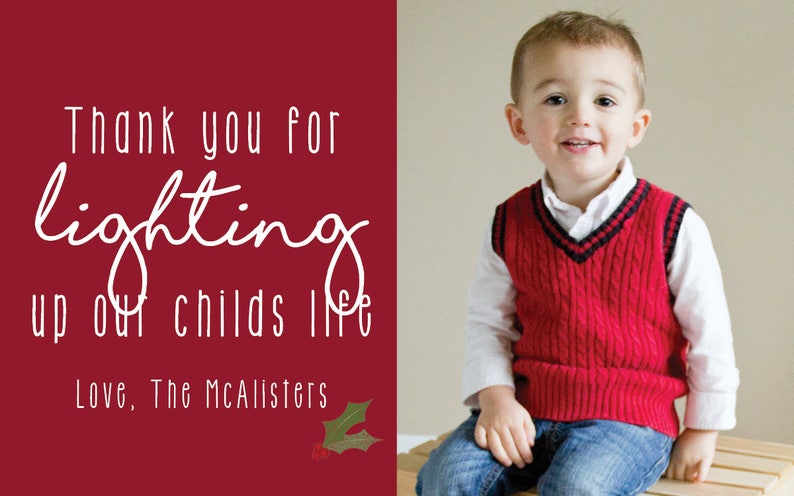 May include: A red Christmas card with white text that reads "Thank you for lighting up our child's life. Love, The McAlister's". A young boy wearing a red sweater vest and blue jeans is sitting on a wooden surface.