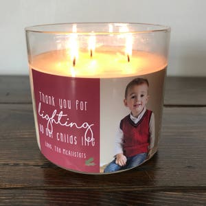 May include: A clear glass candle jar with a red and white label. The label has a photo of a young boy and the text "Thank you for lighting up our child's life. Love, The McAllisters".