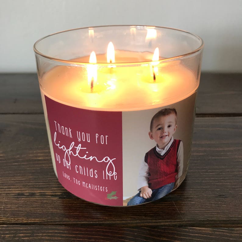 May include: A clear glass candle jar with a red and white label. The label has a photo of a young boy and the text "Thank you for lighting up our child's life. Love, The McAllisters". The candle is lit with three wicks.