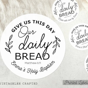 May include: White round labels with black text. The labels read "Give us this day our daily bread" with the Bible verse Matthew 6:11. Some labels say "Emma's Holy Baptism" or "Made with love by Emma". The labels are for a baptism or first communion.