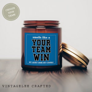 May include: A brown glass candle jar with a gold lid. The label on the jar is blue and says "smells like a YOUR TEAM WIN MY LUCKY GAME DAY CANDLE".
