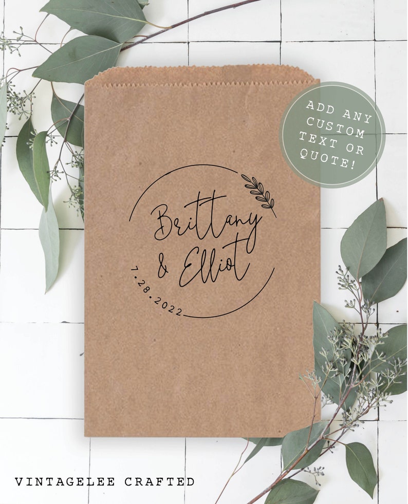 May include: A brown paper bag with a black circle design that says "Brittany & Elliot 7.28.2022". The bag is decorated with a small sprig of greenery. The text "ADD ANY CUSTOM TEXT OR QUOTE!" is written in a circle on the bag.