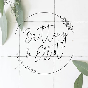 May include: A white background with a black circle containing the text "Brittany & Elliot" and the date "1.28.2022". A small floral design is in the upper right corner of the circle. The text "VINTAGELEE CRAFTED" is at the bottom of the image.