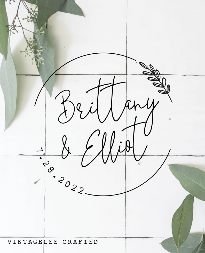 May include: A white background with a black circle containing the text "Brittany & Elliot" and the date "1.28.2022". A small floral design is in the upper right corner of the circle. The text "VINTAGELEE CRAFTED" is at the bottom of the image.