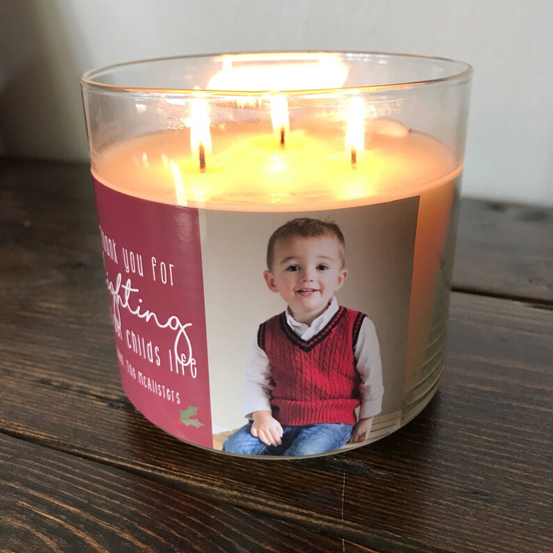 May include: A clear glass candle jar with a red and white label. The label has a photo of a young boy wearing a red sweater vest and blue jeans. The text on the label reads "Thank you for lighting Childs life The McAllisters".
