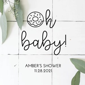 May include: A white tiled background with green eucalyptus leaves on either side. A black and white illustration of a donut with the text "oh baby!"  below it. The text "AMBER'S SHOWER 11.28.2021" is below the illustration. The text "VINTAGELEE CRAFTED" is at the bottom of the image.