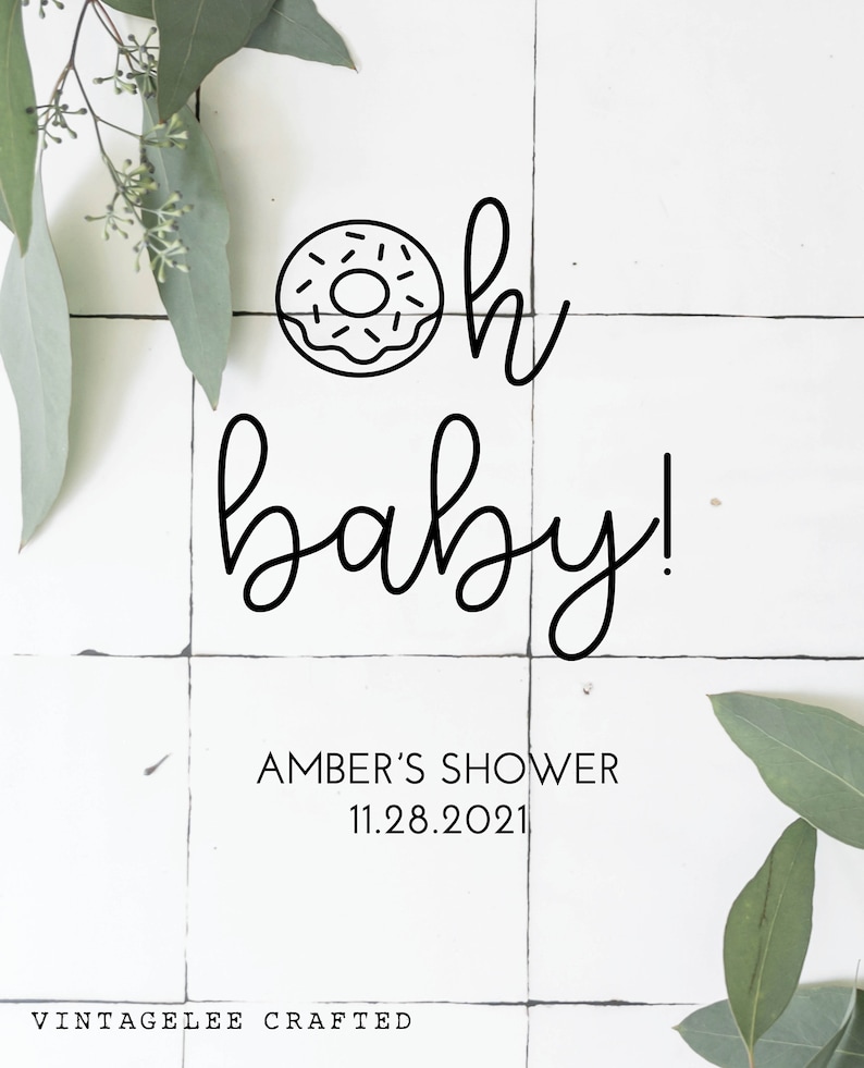 May include: A white tiled background with green eucalyptus leaves on either side. A black and white illustration of a donut with the text "oh baby!"  below it. The text "AMBER'S SHOWER 11.28.2021" is below the illustration. The text "VINTAGELEE CRAFTED" is at the bottom of the image.
