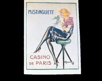 Puede incluir: Cartel vintage con el texto «MISTINGUETT» y «CASINO DE PARIS». La ilustración muestra a una mujer rubia con un loro, sentada en un taburete. El cartel tiene un borde blanco y un estilo art déco clásico.