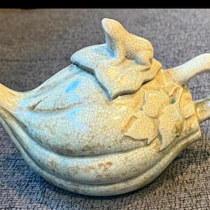 May include: A ceramic teapot with a light blue and beige crackle glaze. The teapot features a leaf-shaped lid and floral detailing on the side. The handle is a loop shape.