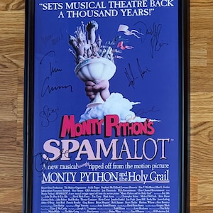 Può includere: Poster incorniciato per il musical "Monty Python's Spamalot" con sfondo blu. L'opera presenta un calice in stile cartone animato tenuto da una mano, con firme e testo tra cui "Un nuovo musical amorevolmente tratto dal film Monty Python and the Holy Grail."