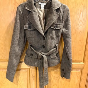 May include: A brown and gray herringbone pattern jacket with a tie belt. The jacket has a notched collar, two front pockets, and button closures. The jacket is hanging on a white hanger.