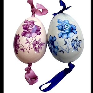 Op de afbeelding: Twee decoratieve eivormige ornamenten. Eén ei heeft een paars rozenontwerp, de andere een blauwe roos. Beide hebben crèmekleurige achtergronden en zijn versierd met bijpassende linten om op te hangen. Ideaal voor Pasen of lente decoratie.