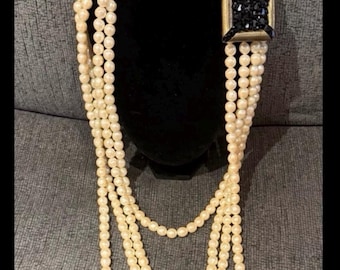 Può includere: Collana di perle a più fili con chiusura rettangolare nera e dorata. La collana è esposta su un busto nero su sfondo grigio. Le perle sono di un colore bianco sporco crema.