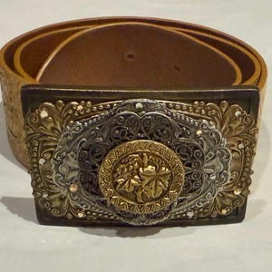 May include: A brown leather belt with a large ornate metal buckle. The buckle is silver and gold with a floral design and a central circular design with a grapevine motif.