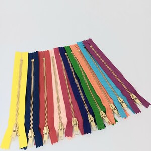 May include: A collection of colorful zippers in various shades, including yellow, navy, red, pink, green, orange, and purple. Each zipper has a gold-colored metal pull and teeth, arranged against a white background. These zippers are ideal for sewing and crafting projects.